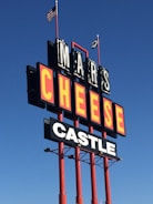 A tall outdoor sign with large bold letters reads 'MARS CHEESE CASTLE'. It is supported by red poles and is set against a clear blue sky. Two flags, including an American flag, are flying at the top.