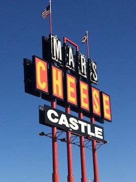 A tall outdoor sign with large bold letters reads 'MARS CHEESE CASTLE'. It is supported by red poles and is set against a clear blue sky. Two flags, including an American flag, are flying at the top.