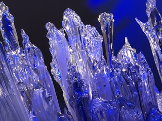 A detailed view of intricately shaped translucent glass-like sculptures with a striking blue illumination. The structures are elongated, resembling icy stalagmites or crystalline formations, reflecting and refracting the surrounding light.