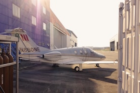 A small jet airplane is parked on an airstrip beside a large hangar. The hangar is painted in bright colors with several windows visible. The airplane is white with a registration number on its tail and has a sleek, modern design. In the foreground, on the left, there are several gas cylinders labeled 'Helium'. The lighting indicates that it is either early morning or late afternoon, casting long shadows.