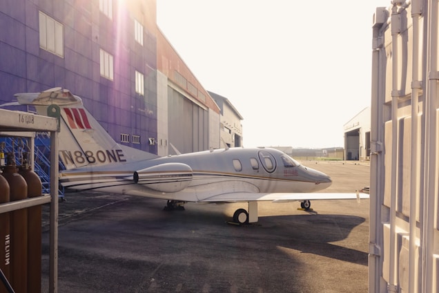 A sleek small airplane parked on a sunlit airfield beside a cozy flight school hangar.