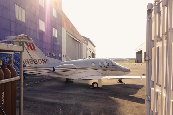 A small jet airplane is parked on an airstrip beside a large hangar. The hangar is painted in bright colors with several windows visible. The airplane is white with a registration number on its tail and has a sleek, modern design. In the foreground, on the left, there are several gas cylinders labeled 'Helium'. The lighting indicates that it is either early morning or late afternoon, casting long shadows.