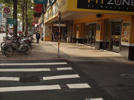 A city street scene with bicycles parked along the sidewalk and a person walking past a gym. The gym entrance is located on the ground floor of a building with bright yellow signage. There are several advertisements on the walls and windows of the gym. On the street, there is a visible pedestrian crossing with white zebra stripes.