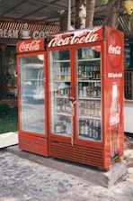 A red Coca-Cola branded refrigerator placed outdoors, filled with various beverages visible through the glass doors. The unit is secured with a chain and padlock. In the background, part of a sign with the words 'CREAM' and 'COFFEE' can be seen. The refrigerator is on a stone or concrete platform, and there are some trees and a green mat nearby.