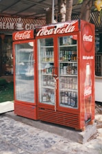 A red Coca-Cola branded refrigerator placed outdoors, filled with various beverages visible through the glass doors. The unit is secured with a chain and padlock. In the background, part of a sign with the words 'CREAM' and 'COFFEE' can be seen. The refrigerator is on a stone or concrete platform, and there are some trees and a green mat nearby.