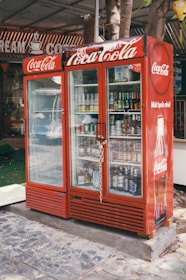 A red Coca-Cola branded refrigerator placed outdoors, filled with various beverages visible through the glass doors. The unit is secured with a chain and padlock. In the background, part of a sign with the words 'CREAM' and 'COFFEE' can be seen. The refrigerator is on a stone or concrete platform, and there are some trees and a green mat nearby.