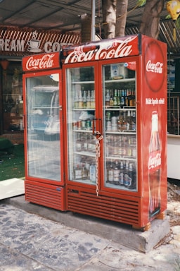 A red Coca-Cola branded refrigerator placed outdoors, filled with various beverages visible through the glass doors. The unit is secured with a chain and padlock. In the background, part of a sign with the words 'CREAM' and 'COFFEE' can be seen. The refrigerator is on a stone or concrete platform, and there are some trees and a green mat nearby.