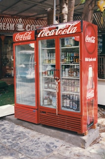 A red Coca-Cola branded refrigerator placed outdoors, filled with various beverages visible through the glass doors. The unit is secured with a chain and padlock. In the background, part of a sign with the words 'CREAM' and 'COFFEE' can be seen. The refrigerator is on a stone or concrete platform, and there are some trees and a green mat nearby.