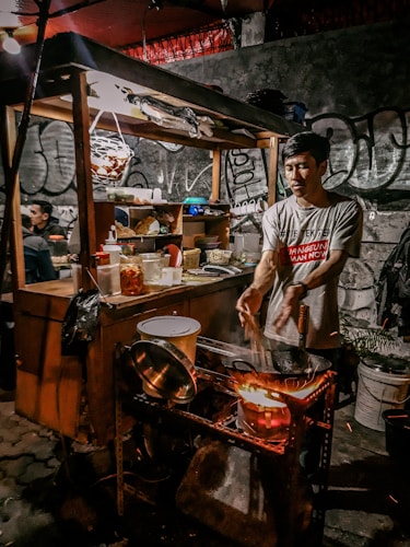 A street vendor in a casual setting, surrounded by cooking equipment and ingredients, prepares food over an open flame in a bustling environment. The vendor is concentrating on stirring the contents in a pot, and the scene is framed by graffiti-covered walls and various kitchen utensils.