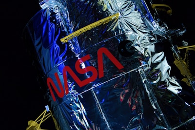 A close-up view of a highly reflective metallic structure, displaying the NASA logo prominently in red. The surface is crinkled, suggesting a foil-like texture, with a blend of silver and blue hues. Yellow structural components are visible, adding contrast to the overall design.