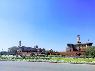 A large, majestic government building featuring colonial architectural style with multiple domes and towers. The structure is made of red and beige stone, surrounded by well-maintained gardens and lush green lawns. The scene is set against a clear blue sky.