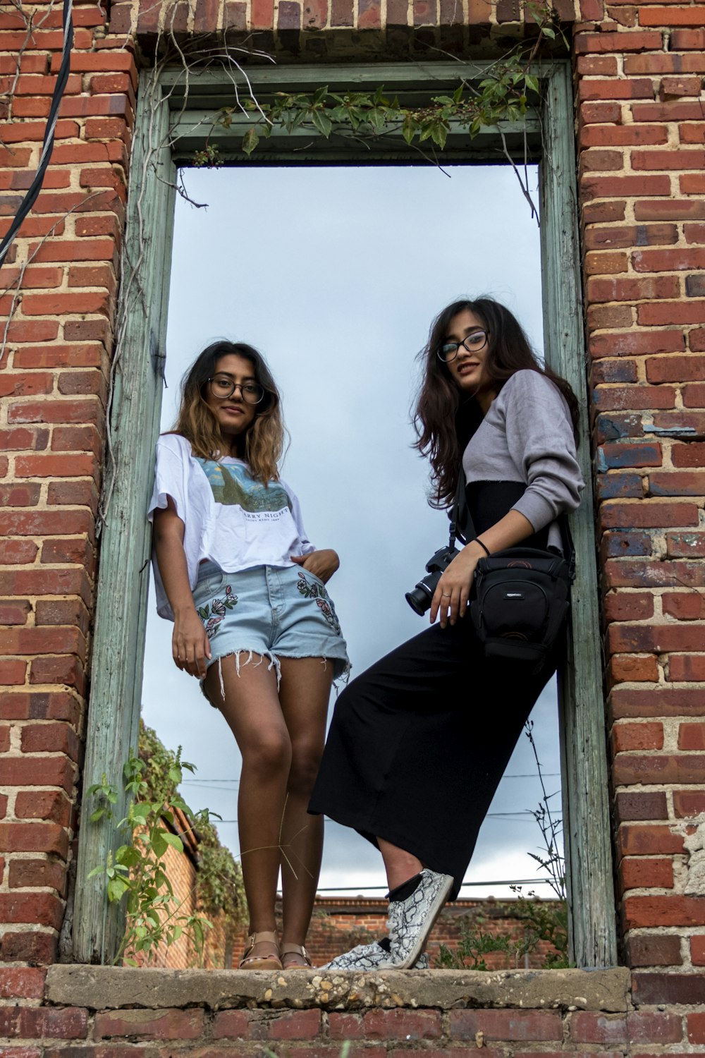 2 women standing beside brown brick wall during daytime