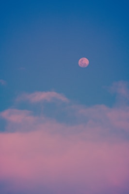 Pink full moon rising behind pink clouds in blue sky
