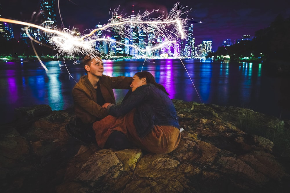 man in black jacket sitting on rock near body of water during nighttime