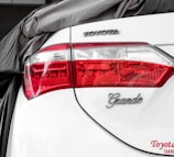 The rear of a white car is partially covered by a gray fabric. The taillight features clear and red sections with intricate detailing. The brand logo 'TOYOTA' and model designation 'Grande' are displayed on the trunk, alongside a visible license plate area.