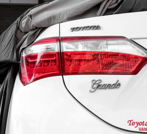 The rear of a white car is partially covered by a gray fabric. The taillight features clear and red sections with intricate detailing. The brand logo 'TOYOTA' and model designation 'Grande' are displayed on the trunk, alongside a visible license plate area.