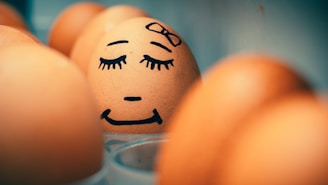 An egg with a drawn face, featuring closed eyes, eyelashes, a smile, and a small bow on its head, surrounded by other plain eggs.
