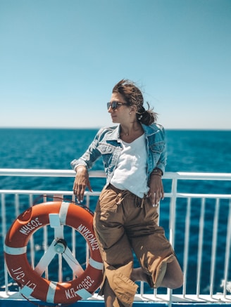 A person wearing sunglasses and a denim jacket leans casually against a ship's railing with a bright orange life preserver marked 'Mykonos' beside them. The background features a vibrant blue ocean under a clear sky.
