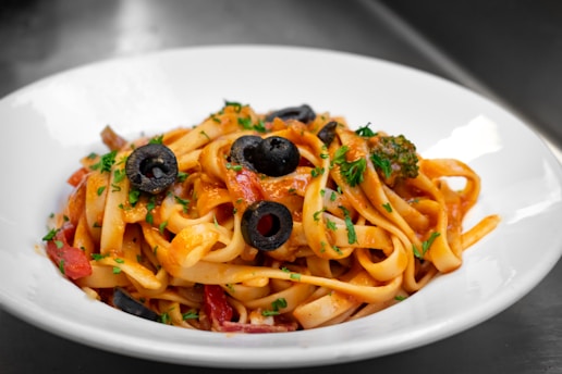 A close-up of a plate containing pasta with a rich tomato sauce. The dish is garnished with slices of black olives and sprinkled with chopped fresh parsley. The pasta is likely fettuccine or tagliatelle, and the sauce appears to include pieces of vegetables, possibly bell peppers and broccoli.