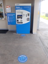 A blue and white ticket vending machine is situated against a wall with various notices around it, including a sign about coronavirus safety measures. In front of the machine, the floor has circular markers indicating where to stand to maintain social distancing. The setting appears to be a transit station with a customer service window nearby.