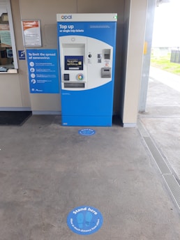 A blue and white ticket vending machine is situated against a wall with various notices around it, including a sign about coronavirus safety measures. In front of the machine, the floor has circular markers indicating where to stand to maintain social distancing. The setting appears to be a transit station with a customer service window nearby.