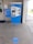 A blue and white ticket vending machine is situated against a wall with various notices around it, including a sign about coronavirus safety measures. In front of the machine, the floor has circular markers indicating where to stand to maintain social distancing. The setting appears to be a transit station with a customer service window nearby.
