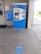 A blue and white ticket vending machine is situated against a wall with various notices around it, including a sign about coronavirus safety measures. In front of the machine, the floor has circular markers indicating where to stand to maintain social distancing. The setting appears to be a transit station with a customer service window nearby.