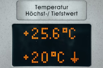 A digital temperature display board showing current and maximum temperature readings. The current temperature is 25.6 degreeand another value of 20 degrees Celsius with a downward arrow is shown. The board features orange LED numbers on a black background with a metal sign above in a different language indicating temperature-related information.
