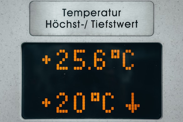 A digital temperature display board showing current and maximum temperature readings. The current temperature is 25.6 degrees Celsius, and another value of 20 degrees Celsius with a downward arrow is shown. The board features orange LED numbers on a black background with a metal sign above in a different language indicating temperature-related information.