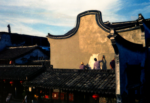 Several individuals are engaged in the process of painting or plastering a wall of a traditional Asian building with a distinctive curved roof. The setting appears to be late in the day with a soft glow from the ambient lighting. Shadows are visible due to the angle of the sunlight.
