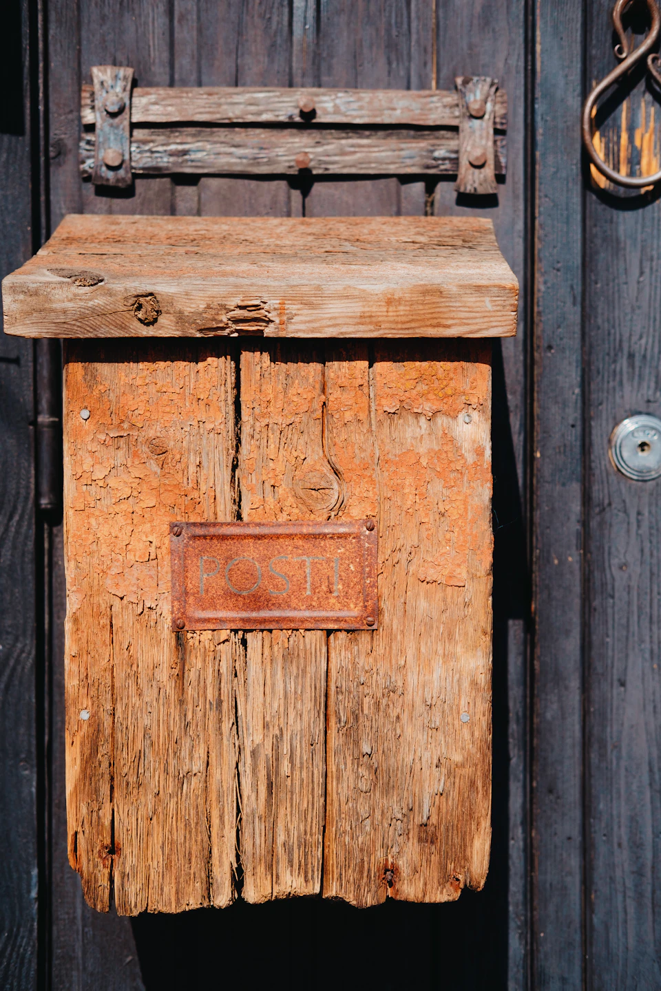 The image shows a wooden mailbox with a rustic charm, painted in shades of brown and adorned with a weathered sign that reads "RUSTIC". The mailbox is situated on a dark wooden door, which has a metal handle for opening it. A small window can be seen on the right side of the door.