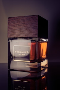 A sleek perfume bottle with a wooden cap is prominently displayed. The label reads 'Imperial Oud for Men'. The design combines glass and wood, giving it an elegant and masculine appearance. The lighting highlights the rich amber liquid inside.