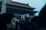 A crowd of people is gathered in front of a historic building with traditional Chinese architecture, focusing their attention on capturing photos and videos with their smartphones. A prominent portrait of a figure is displayed on the building, contributing to the significance of the moment.