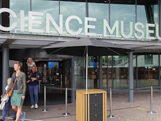 Entrance to a modern building with large glass windows and a sign that reads 'Science Museum'. People are entering and exiting the building, including a woman with two children.
