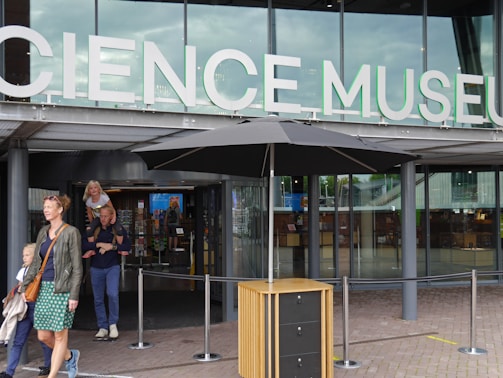 Entrance to a modern building with large glass windows and a sign that reads 'Science Museum'. People are entering and exiting the building, including a woman with two children.