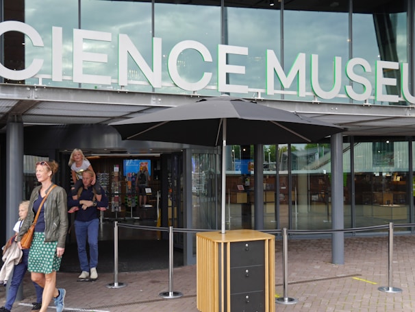 Entrance to a modern building with large glass windows and a sign that reads 'Science Museum'. People are entering and exiting the building, including a woman with two children.