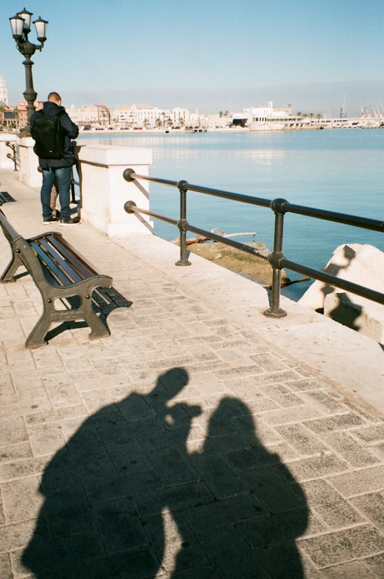 man in black jacket standing on gray concrete floor near body of water during daytime in Bari Italy