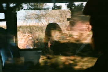 A reflective window captures the blurred image of two people inside a train or bus, with one holding a camera. The exterior landscape is visible, showing trees and the warm tones of an outdoor environment.