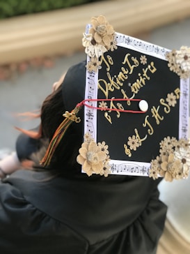 A close-up view of a decorated graduation cap adorned with beige flowers and gold lettering that reads 'To define is to limit' surrounded by a musical note border. The cap is seen from above, worn by a person in a black graduation gown.