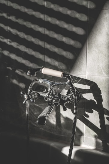 A vintage-style bathtub faucet with a handheld shower attachment is mounted on a tiled wall. The metal fixtures have a polished chrome finish, and light creates striped shadows across the tiles, adding a dramatic effect.
