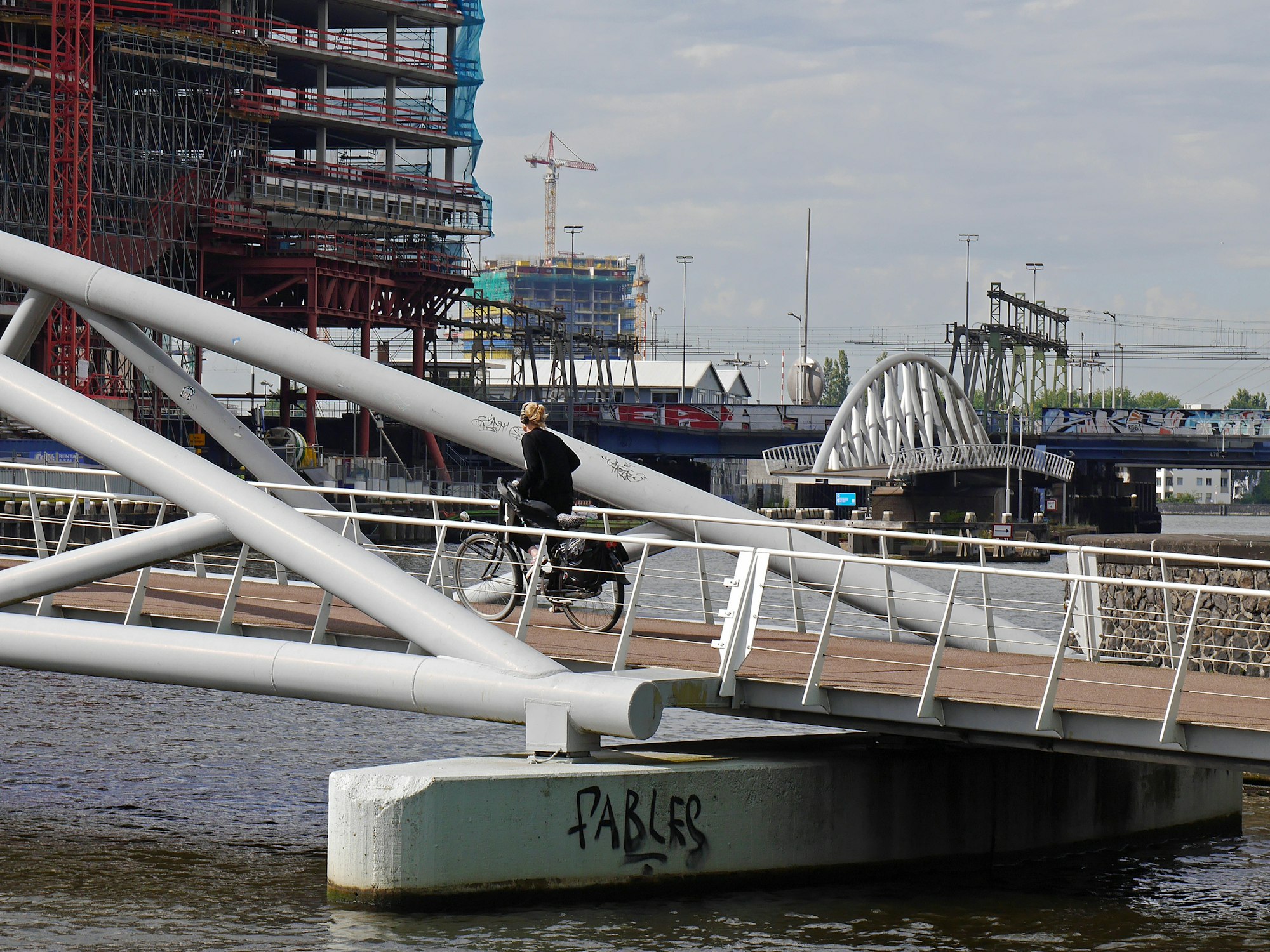 A woman is driving up the bridge, close to the NEMO museum in city center of Amsterdam. Picture of modern bridge architecture. Free urban photo of Fons Heijnsbroek, July 2020, The Netherlands. 

Nederlands: een vrouw fiets de brug bij het NEMO museum op, op het Oosterdok van Amsterdam. Links is de nieuwbouw van Booking.com op het puntje van het Oosterdokseiland. Gratis foto van moderne brug architectuur, juli 2020, Nederland.  