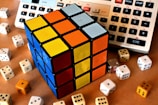 A Rubik's Cube with sides showing blue, yellow, orange, and white is placed among several six-sided dice of varying colors. In the background, parts of two electronic calculators are visible.