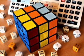 A Rubik's Cube with sides showing blue, yellow, orange, and white is placed among several six-sided dice of varying colors. In the background, parts of two electronic calculators are visible.