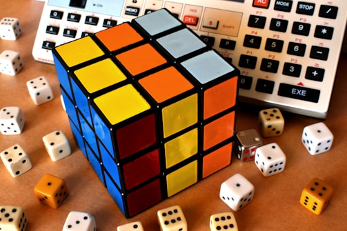 A Rubik's Cube with sides showing blue, yellow, orange, and white is placed among several six-sided dice of varying colors. In the background, parts of two electronic calculators are visible.