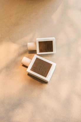 Two perfume bottles are lying on a light surface, casting soft shadows. The bottles are white with minimalistic, brown labels.