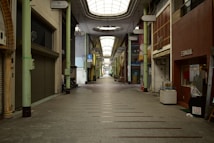 A long, narrow indoor shopping street with a high ceiling and soft natural lighting. The shops on either side have closed shutters, and there is minimal foot traffic. Signs in different languages hang above the entrances, and a few bicycles are parked along the sides.