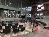 A large industrial-style room with vintage control panels and gauges surrounds a semi-circle of empty chairs arranged for an event or meeting. A camera on a tripod is set up, indicating filming or broadcasting. The architecture features large metal-framed windows and visible ceiling beams.