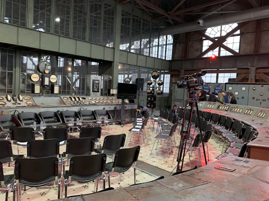 A large industrial-style room with vintage control panels and gauges surrounds a semi-circle of empty chairs arranged for an event or meeting. A camera on a tripod is set up, indicating filming or broadcasting. The architecture features large metal-framed windows and visible ceiling beams.