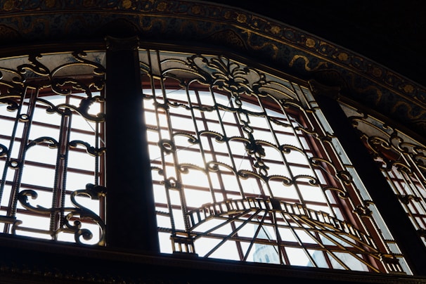 Intricate wrought iron patterns cover a large window, allowing sunlight to filter through and cast shadows on the ornate designs. The window's frame is detailed with elaborate scrollwork and embellishments, surrounded by richly decorated moldings.