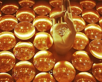 A golden hand positioned in a gesture commonly associated with meditation, surrounded by numerous shiny circular discs that create a textured backdrop. The discs reflect light, contributing to a luminous and harmonious visual effect.